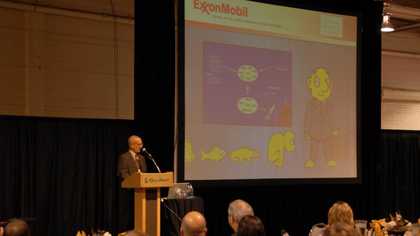 A man in a suit speaking at a lectern in front of an audience, with a presentation visible on a large projector screen behind him.