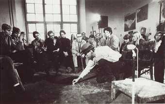 Black-and-white photograph in an art studio. Joseph Beuys sits on a chair and bends to draw in chalk on the floor. An audience sits around and watches.