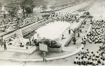 Black-and-white photograph looking down on a landscaped park and rectangular pond around which groups of people are gathered, including students in uniform