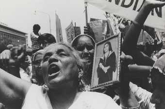 A large crowd of figures shout and hold up photographs and banners in an outdoor setting.