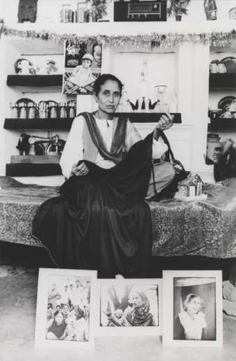 A figure sits on a bed holding a dark swathe of cloth, with three photographs displayed at their feet.