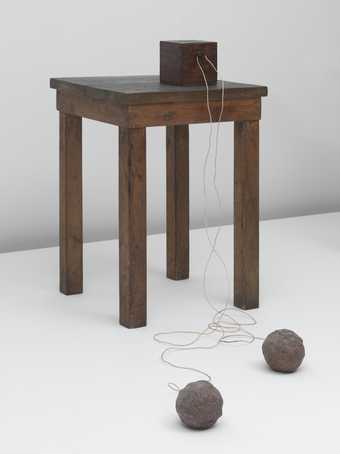 Installation featuring an accumulator – a kind of rechargeable battery in which energy can be stored – sits on top of a small wooden table and is attached by wires to two balls of clay on the floor