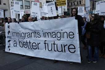 A group of people standing in a street holding placards and a large banner that reads ‘Immigrants imagine a better future’.