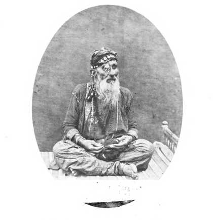 A figure with a long beard sits cross-legged on a bed, wearing a decorated hat and eye patch.