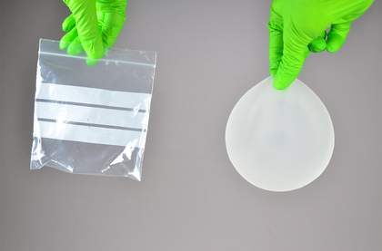Two hands in green gloves against a grey background, one holding an empty, transparent ziplock bag, and the other holding a semi-transparent breast implant.