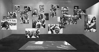 Photographs of people sitting, shouting, giving speeches and protesting hang in a gallery at various heights, with another image and some text projected on the floor beneath them.