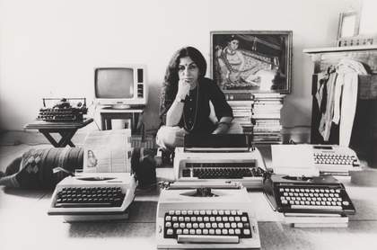 A figure sits cross-legged on the floor in an indoor setting, their chin resting on their hand, surrounded by typewriters, a television, a painting of a seated figure and a chest of drawers with cloth spilling out of it.