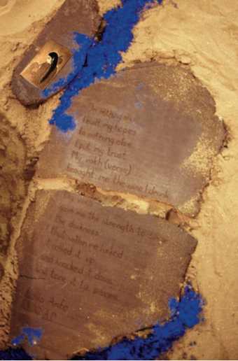 A stone tablet with writing carved into it, surrounded by patches of sand and dry pigment in yellow and blue.