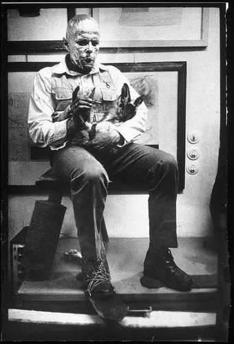 Black-and-white photograph of Joseph Beuys performing How to Explain Pictures to a Dead Hare. He sits in front of a wall of framed pictures, his face covered in gold leaf, a dead hare cradled in his arms, his right foot tied with string to an iron sole