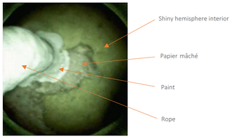 A blurred image showing a circular greenish brown area with lumps of brown papier mâché at its centre, a lumpy section of white paint to the left of the papier mâché, and on far the left, very close to the camera lens, a thick white tube-shaped piece of r