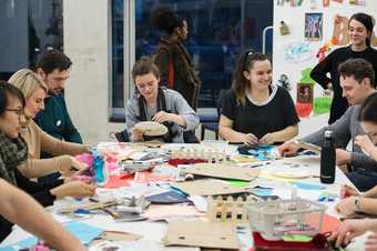 A group of people sit around a table making objects using the craft materials that are in front of them.