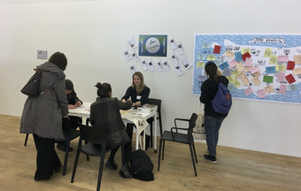 A small group of people sit writing and talking at a table, while another person looks at a large wall map with coloured pieces of paper pinned to it.
