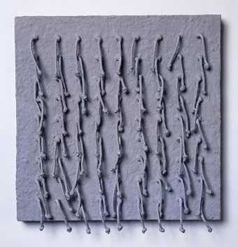 A grey, square-shaped sculpture with eighty-one short pieces of cord with knots at their ends attached to it and hanging downwards in evenly spaced rows.