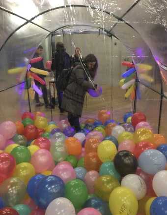 A view from inside a clear, tunnel-like structure, the lower half of which is filled with balloons, with a person standing at the entrance of the tunnel holding a balloon.