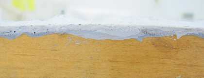 A horizontal view of the edge of a piece of wood with a thin layer of darker grey paint on it and a layer of lighter grey paint over the top.