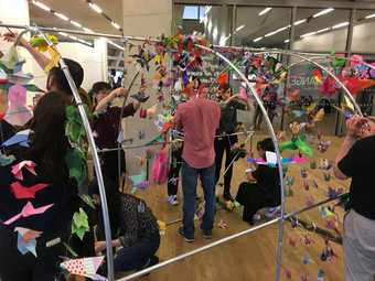 A group of people sit talking and working around a table, framed by an installation of hand-made flowers.