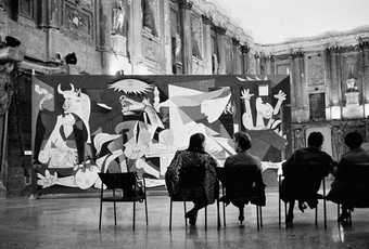 A black and white photograph of Picasso’s painting Guernica installed within a large old Italian building, with four individuals sitting on chairs a short distance in front of the painting.