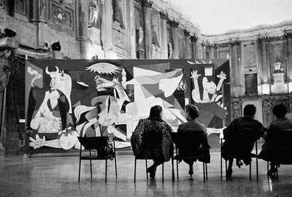 A black and white photograph of Picasso’s painting Guernica installed within a large old Italian building, with four individuals sitting on chairs a short distance in front of the painting.