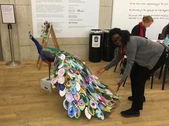 A person faces the camera and presents a large sculpture of a peacock with an array of brightly coloured feathers.