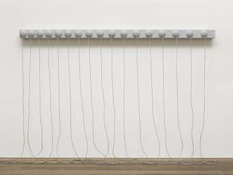 A grey sculpture consisting of a narrow, horizontal panel with seventeen hemispheres on its face, fixed to the wall around two metres above the floor, with long ropes from the centre of each hemisphere that hang down and rest in coils on the floor