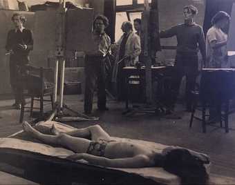 A black and white photograph of a studio where students standing at easels paint a model wearing underwear and lying on a couch in the foreground. Bomberg is in conversation with a student, pointing at their easel.