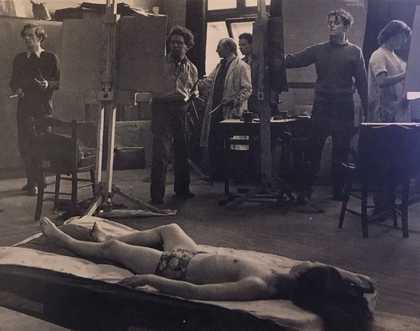 A black and white photograph of a studio where students standing at easels paint a model wearing underwear and lying on a couch in the foreground. Bomberg is in conversation with a student, pointing at their easel.