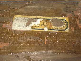 Photograph of a partially ripped yellowed label with a black border under a coat of cracking varnish.