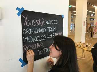 A person writes on a blackboard with coloured chalk. The text reads ‘Youssra, who is originally from Morocco, uses thyme rosemary…’