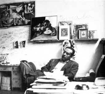 A black and white photograph of a man reclining in a chair, with books and papers in front of him and on the wall behind him a poster of Picasso’s painting Guernica, a painting of a man eating spaghetti, and various black and white portrait photographs.