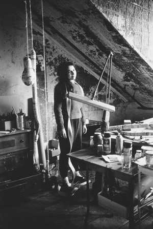 A black and white photograph of the artist in a bare-walled studio space, with rounded objects hanging from the ceiling on ropes to her right, and an array of long sculptural forms, bottles and glasses on a table in front of her.
