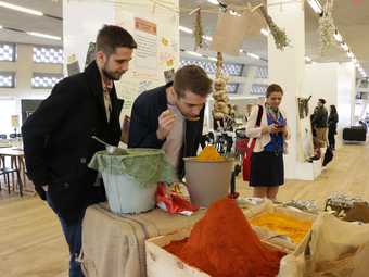 Two people stand behind a table covered with mounds of spices, with one of the people lifting a spoon of yellow spice to their nose to smell it.