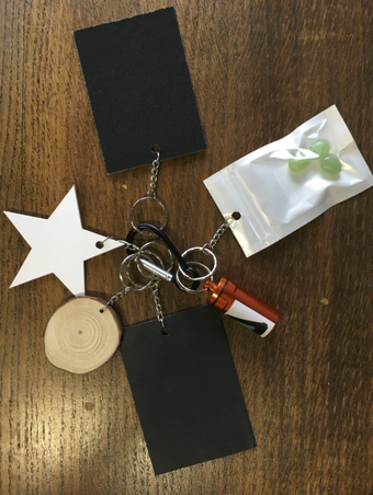 A key chain featuring two black rectangles, a white star, a circle of wood, a packet with three small green pebble-like objects in it, and a miniature red bottle.