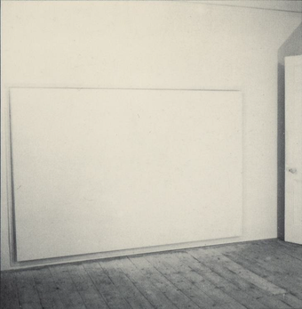 A photograph taken at an angle of a large white canvas hanging just off the floor in a room with a wooden floor.