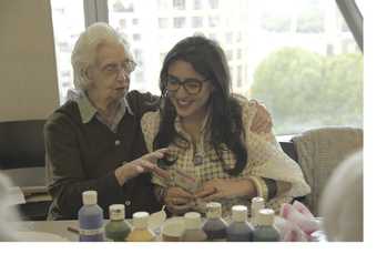 An older person and a younger person sit side by side talking and smiling, with an array of tubes of paint in front of them. The older person has their arm around the younger and is gesticulating as if explaining something.