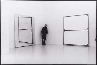 A photograph of Uglow leaning against a wall in a corner of a gallery, between two works.