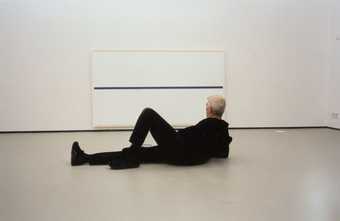 A photograph of Uglow reclining in a gallery, looking at a canvas with a single horizontal line across its middle.
