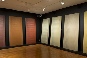 An installation photograph of tall paintings with increasing numbers of vertical waves arranged in the corner of a gallery.