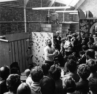 Black-and-white photograph of Tom Hudson speaking in front of a painting in a room full of standing students.