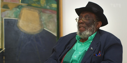 Portrait of Frank Bowling sitting in a chair in front of a painting