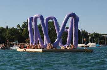 Purple Franz West sculpture positioned by a lake with bathers sitting within it