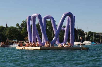 Purple Franz West sculpture positioned by a lake with bathers sitting within it