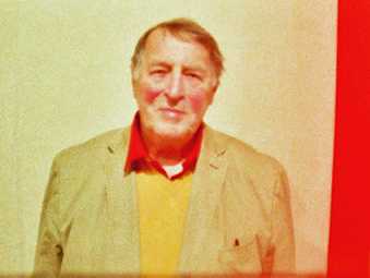 Portrait of the artist Franz Erhard Walther. He wears a red shirt with a yellow jumper. On the wall behind him is a bright stripe of red paint