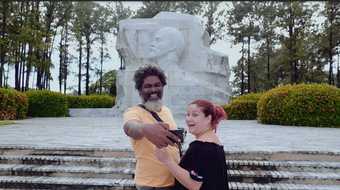 film still of two adults taking a selfie in front of a monument in Cuba