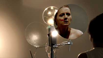Film still shot from behind as a woman looked in one of three round mirrors. She has a wet red line from her nose to her cheek which could be lipstick, paint or blood.