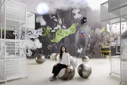 artist Haegue Yang sits on a metal circular ball in a room that has lots of chalk drawings on the wall. They have an abstract shape painted on their face.