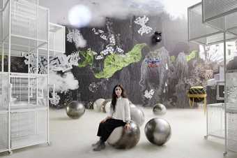 artist Haegue Yang sits on a metal circular ball in a room that has lots of chalk drawings on the wall. They have an abstract shape painted on their face.