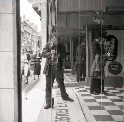 Black and white photograph of Nigel Henderson taking a picture of himself in a mirrored wall by a busy street