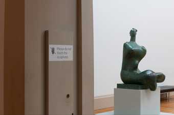 a 'do not touch the sculpture' sign appears next to green bronze sculpture of a person sitting on a plinth
