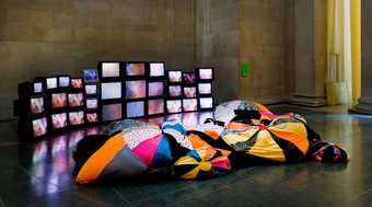 two people are dressed in multicoloured fabric, on the floor of Tate Britain gallery with a series of TVs on the wall behind