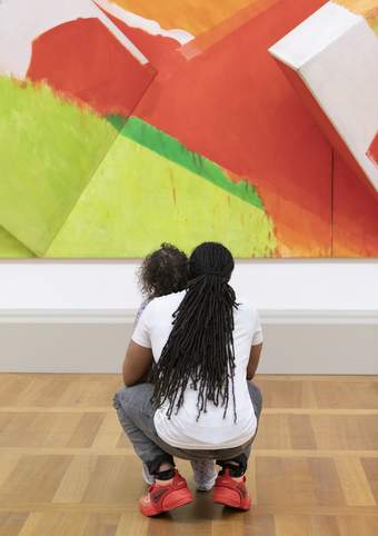A parent crouching down with their arms around their child looking at an artwork titled Green and White 1969 by artist Sandra Blow.